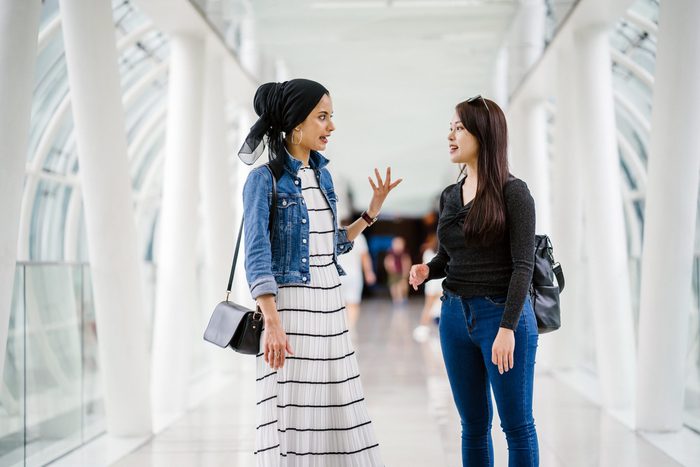 A portrait of two friends standing on a modern, white overpass. One is a Muslim woman and the other is Chinese. They are both young, attractive and happy as they stand next to one another. 