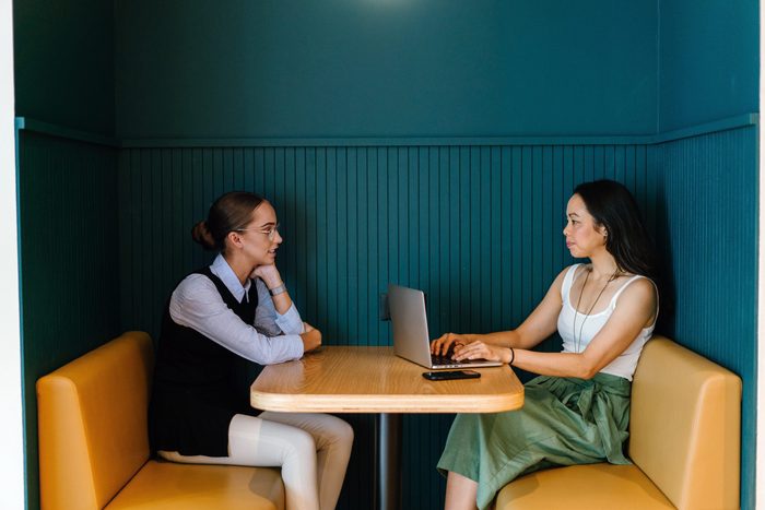 rude conversation habits - A Caucasian and Asian woman sit in a trendy booth in an office and have a business discussion in the office. They are both young, attractive and are having a focused talk about work. 