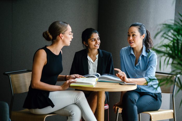A young Indian Asian woman is having a casual business meeting with her team in a meeting room. They are sitting and having an animated conversation. The team is diverse (Caucasian and Chinese woman).