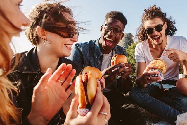 Group of friends sitting on mountain top eating burger. Excited young men and women enjoying and partying outdoors.