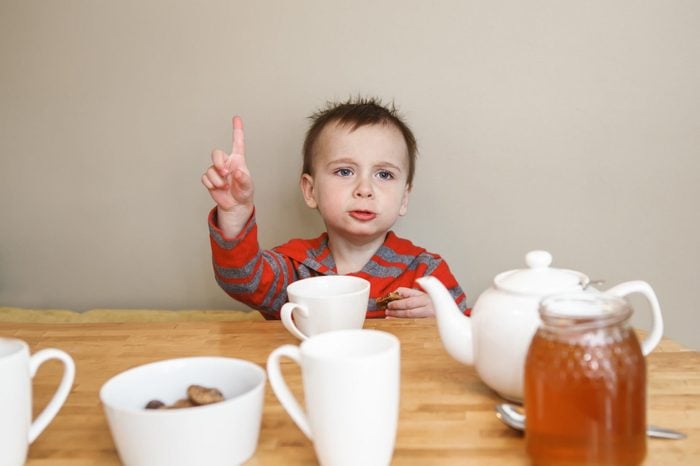 Close-up candid natural portrait of cute little boy toddler in kitchen drinking tea juice, making funny face, showing finger, lifestyle documentary style, grainy film effect