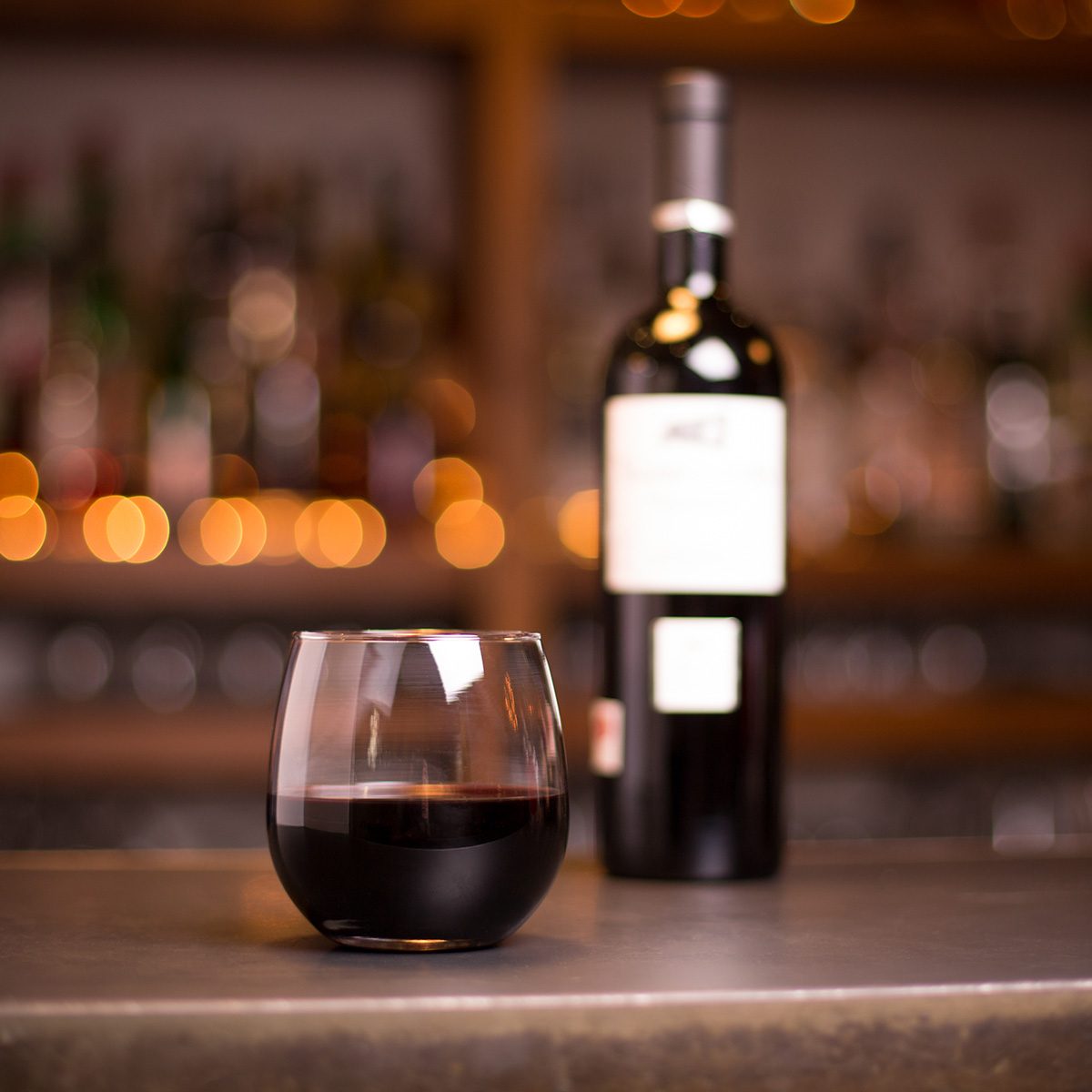 Low angle close up perspective of crystal clear stemless wine glass with traditional round goblet shape filled with rich red wine on metal counter top bar with blurry bottle and restaurant background
