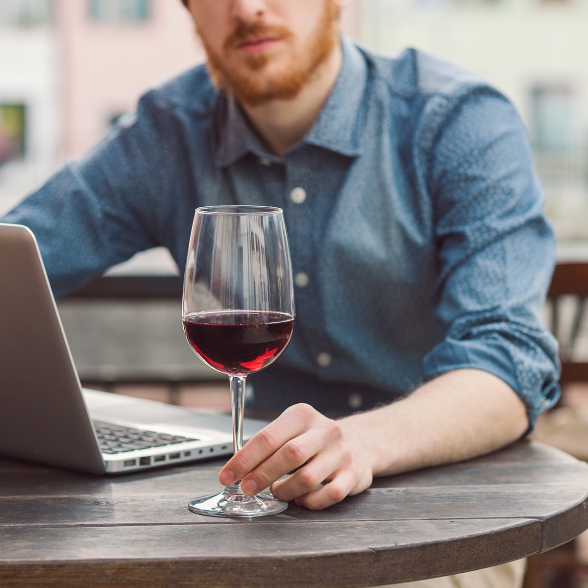 Young hipster man sitting at outdoor bar table with his laptop and having a glass of wine