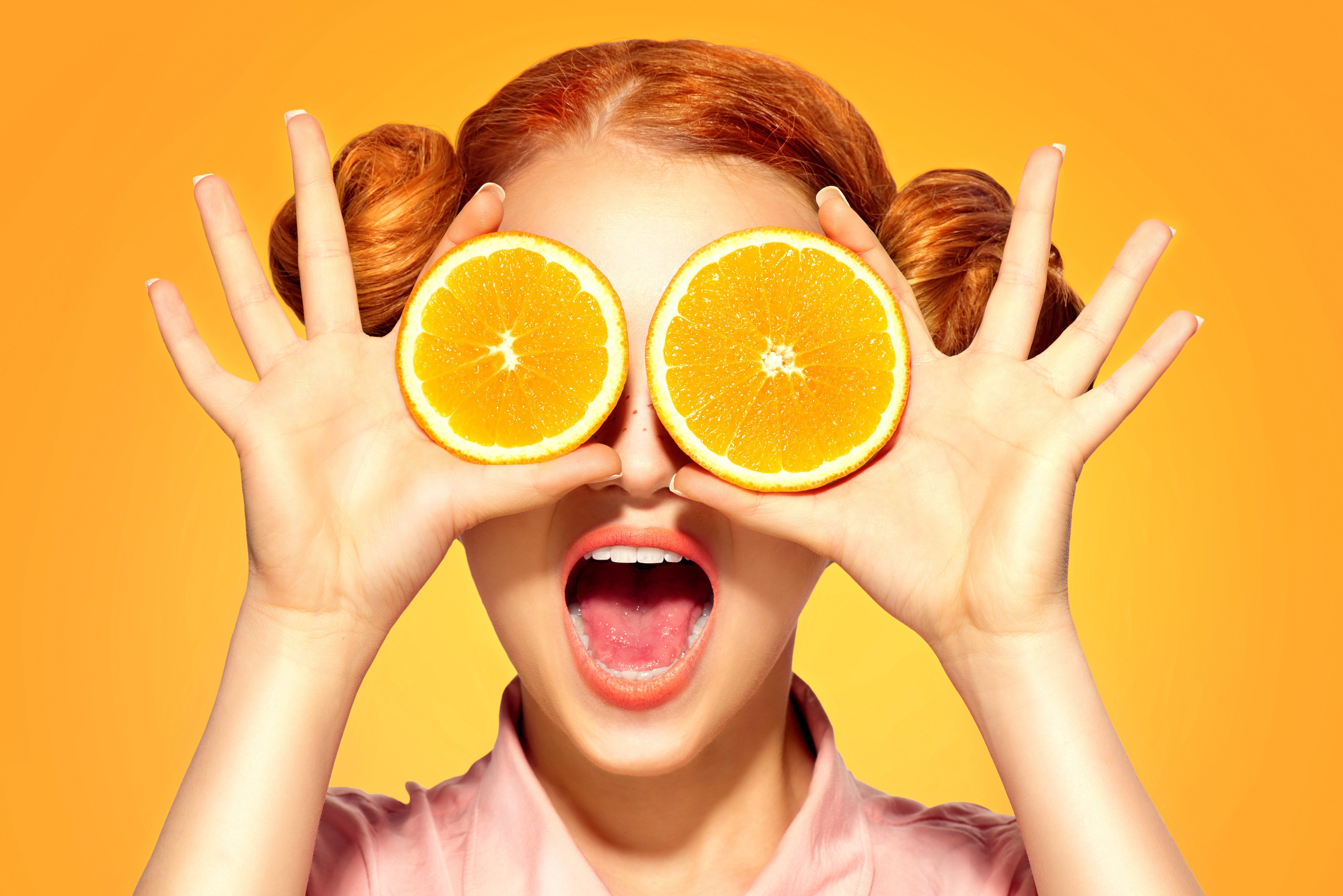 Beauty Model Girl takes Juicy Oranges. Beautiful Joyful teen girl with freckles, funny red hairstyle and yellow makeup. Professional make up. Holding Orange Slices and laughing, emotions