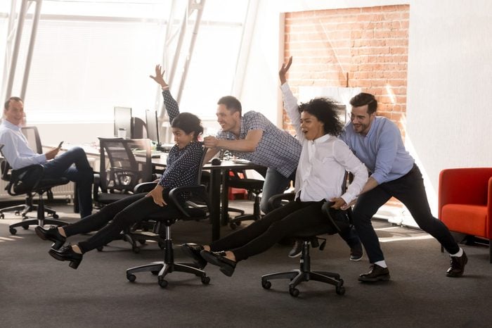 Carefree excited diverse workers having fun riding oh chairs celebrating friday together, happy employees enjoy funny competition laughing together feel great at work break, friendly office team game