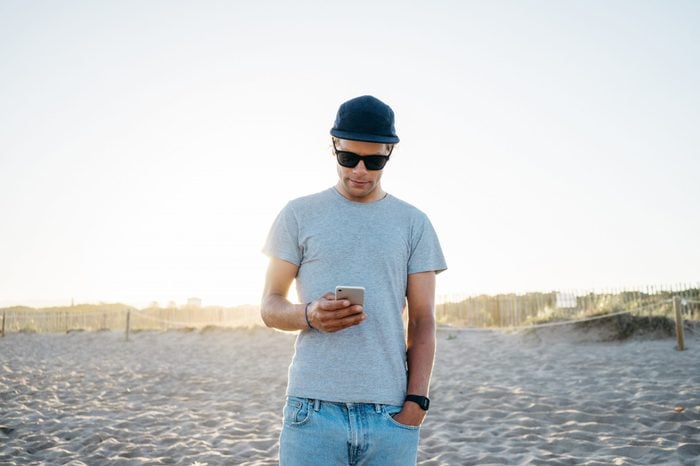 fit young attractive man in a gray casual t-shirt holds mobile phone. picture of male using white smartphone on the beach during beautiful sun light at the holidays. close up of a person touch screen 