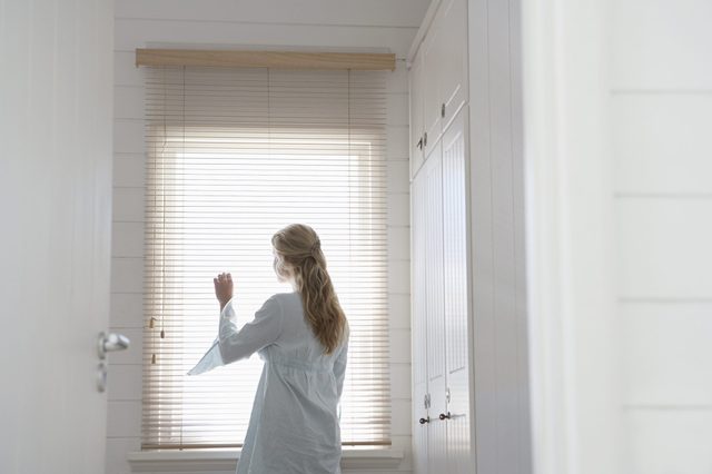 Rear view of young woman looking through window blinds at home