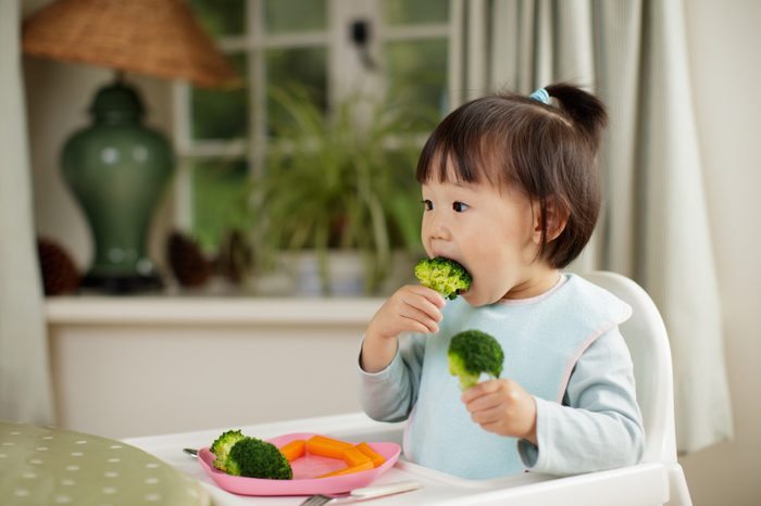 toddler girl eating healthy vegetable sitting on high chair beside a dinner table at home