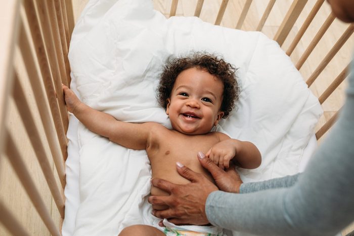 Happy baby boy in a crib. Cropped hands of mother tickling son lying on bed at home.