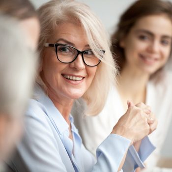 Smiling aged businesswoman in glasses looking at colleague at team meeting, happy attentive female team leader listening to new project idea, coach mentor teacher excited by interesting discussion