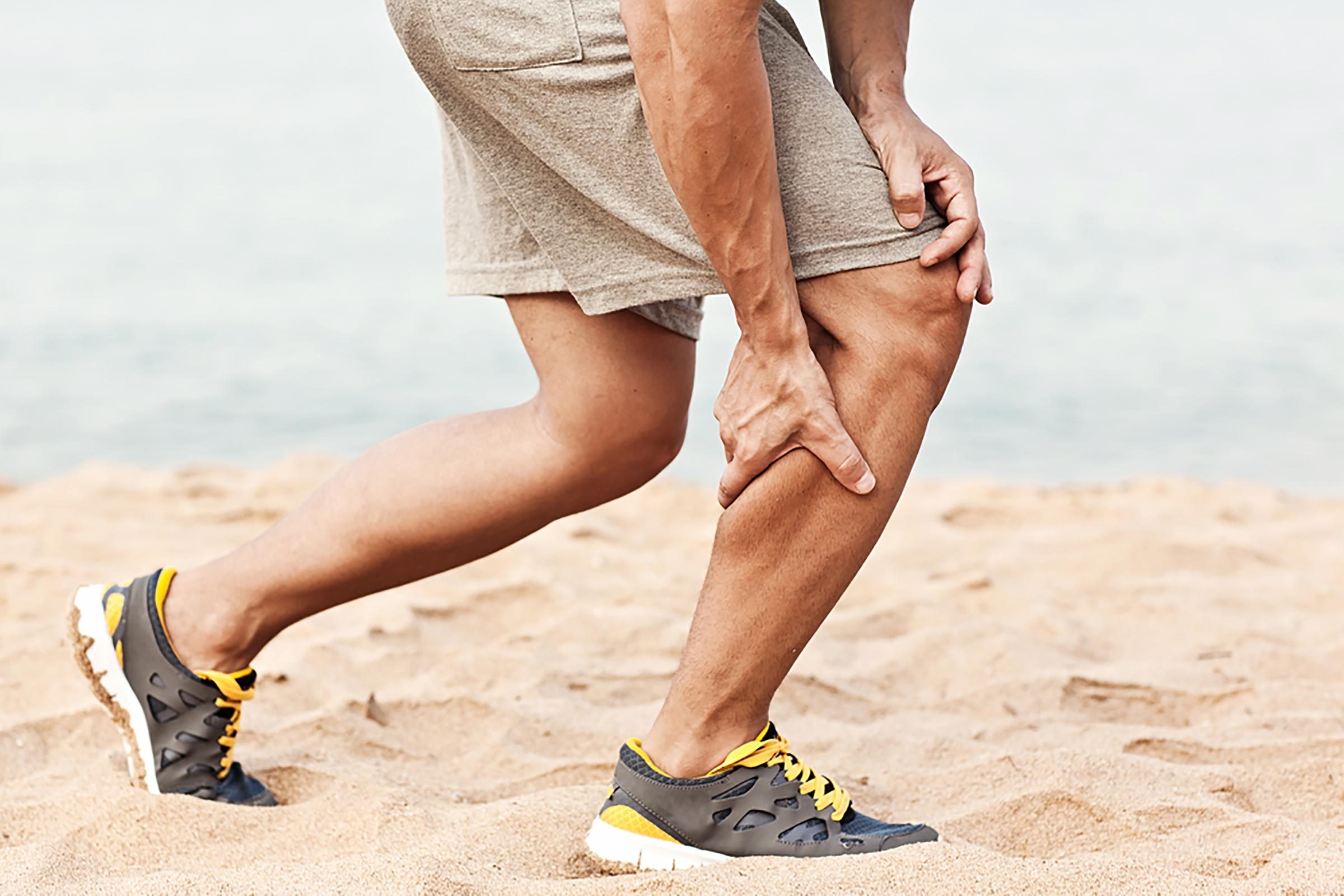 Man on the beach in running shoes, grabbing his calf muscle