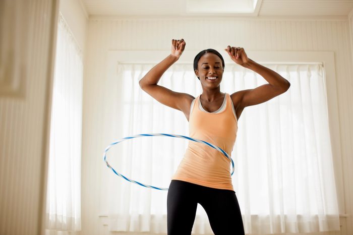 Happy young woman hula hooping in her living room.