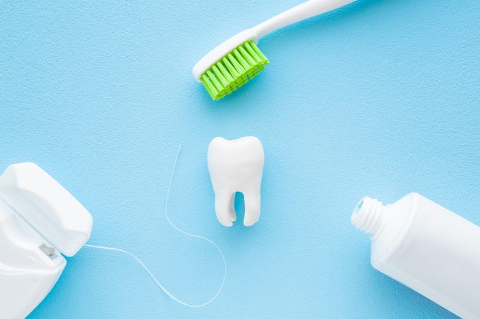 Tube of toothpaste, toothbrush with green bristle and container of dental floss around white tooth on pastel blue background. People teeth hygiene. Closeup.