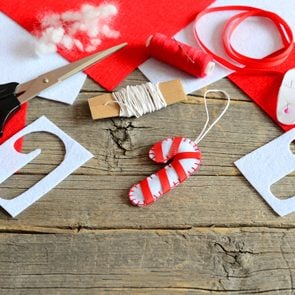 Felt Christmas candy cane ornament, scissors, paper template, thread, needle, red and white felt pieces and scraps, filler, ribbon on old wooden background. Christmas DIY concept. Kids holiday crafts