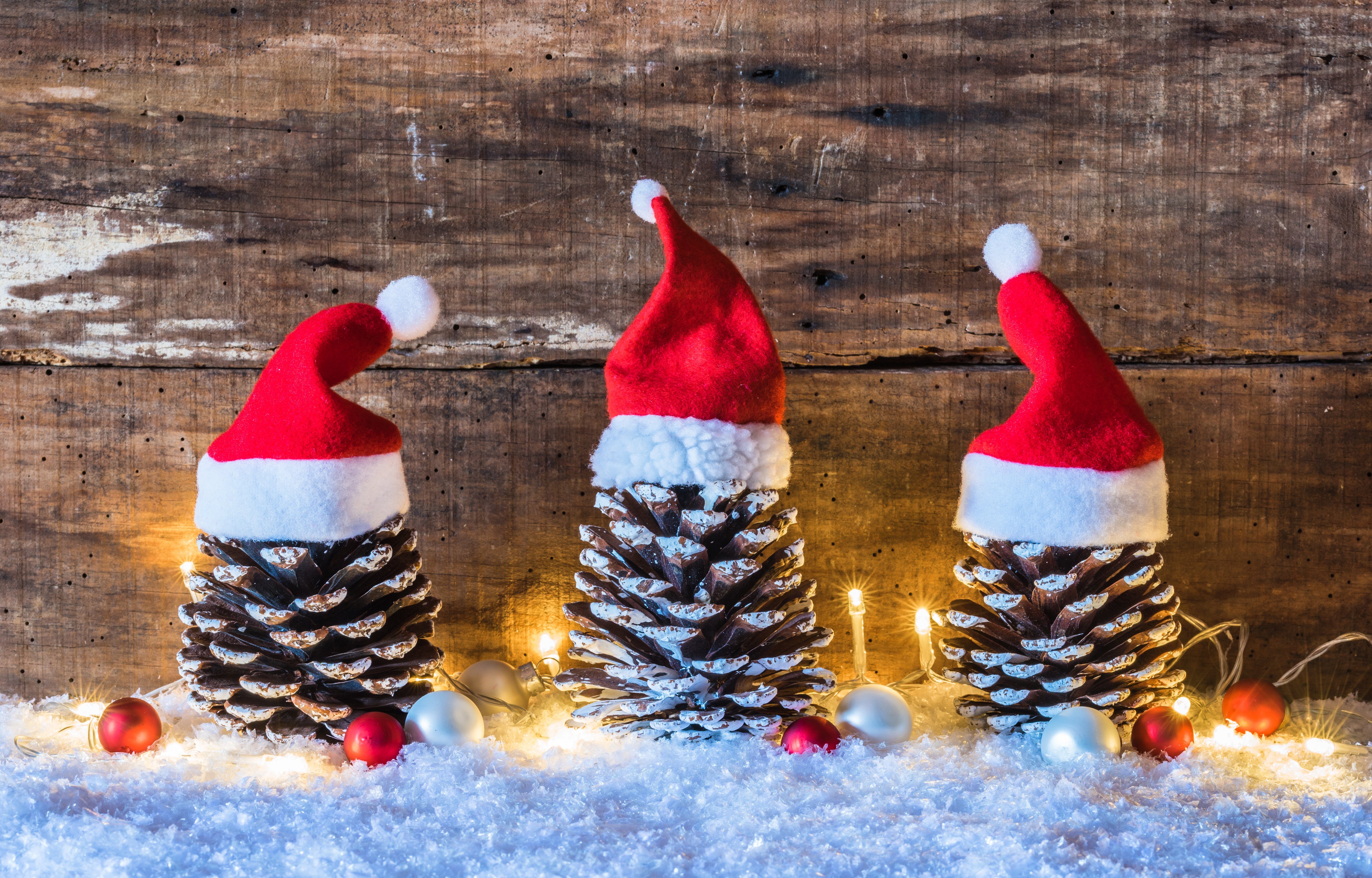 Christmas season, rustic decoration, Santa hats on pine cones with lights at snow and brown wooden background.