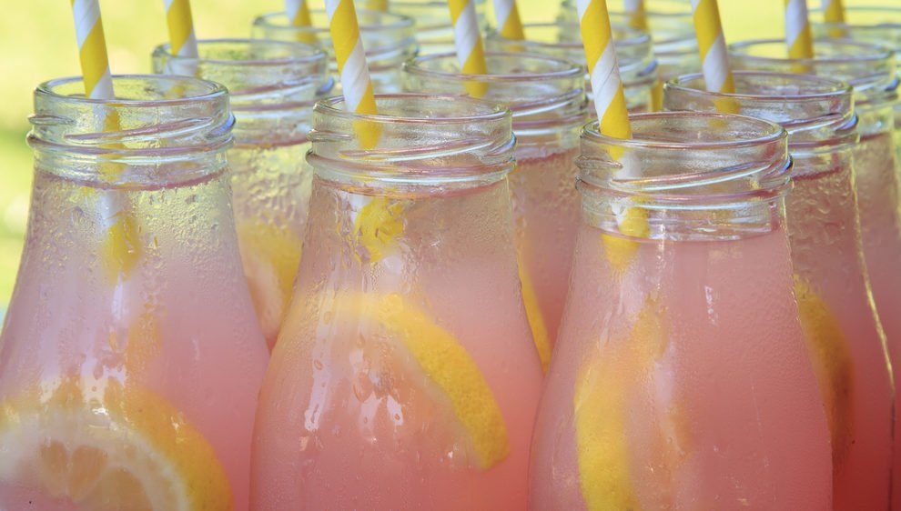 Close up of picnic party in the park drink table with large pitcher and tops of glass bottles filled with ice cold pink lemonade and fresh lemons with yellow swirled straws