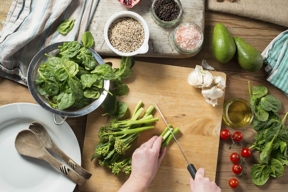Overhead shot of womans hands chopping Broccoli for a salad with ingredients including spinach, tomatoes, quinoa, salt and pepper, garlic, avocado, olive oil, onions and pomegranate.