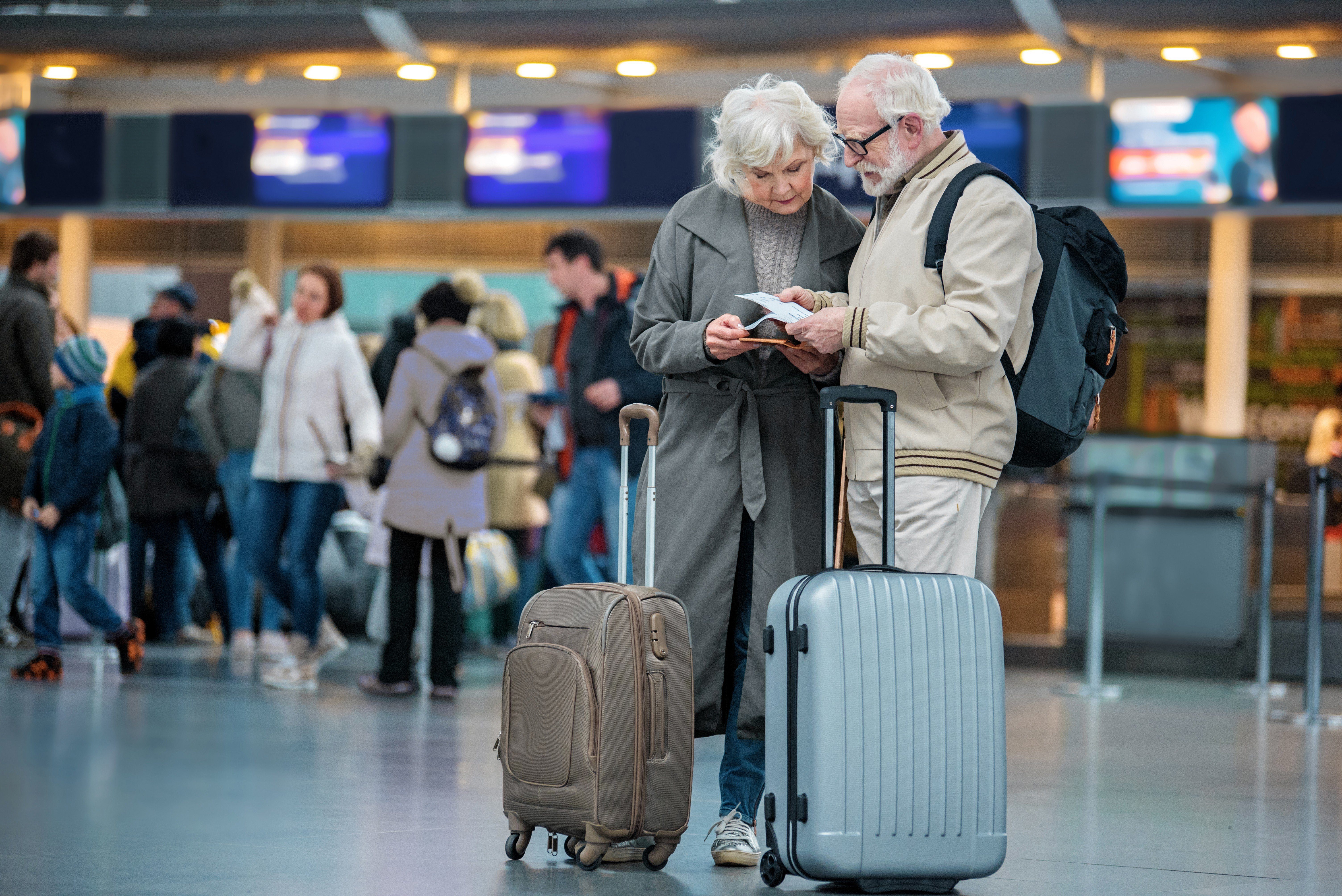 Checking trip details. Full length of serious senior wife and husband are standing with suitcases at international airport and looking at flight tickets with concentration. Copy space in left side