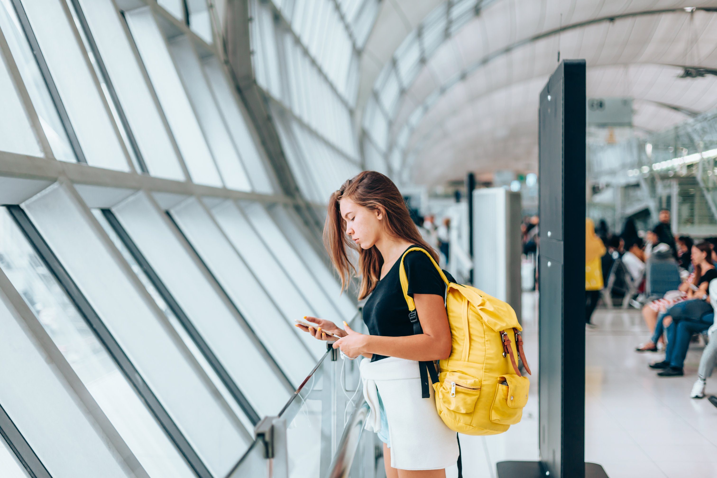 Teen girl using smarthphone while waiting for international flight in airport departure terminal. Young passenger with backpack travelling on airplane. Teenager tourizm abroad alone concept.