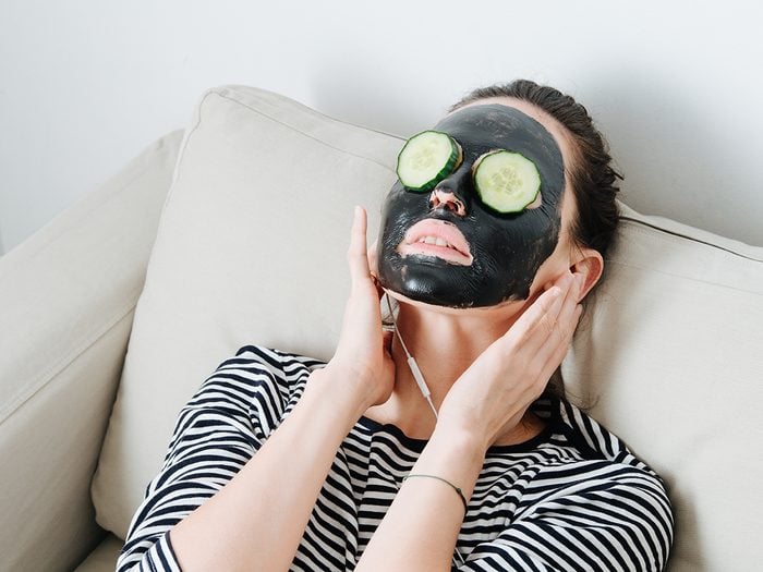 What does activated charcoal do - Young woman with bamboo charcoal peel off facial mask. Sitting in a couch at home, leaning her back. Hair in a bun. Cucumber slices over her eyes. Listening to the music.