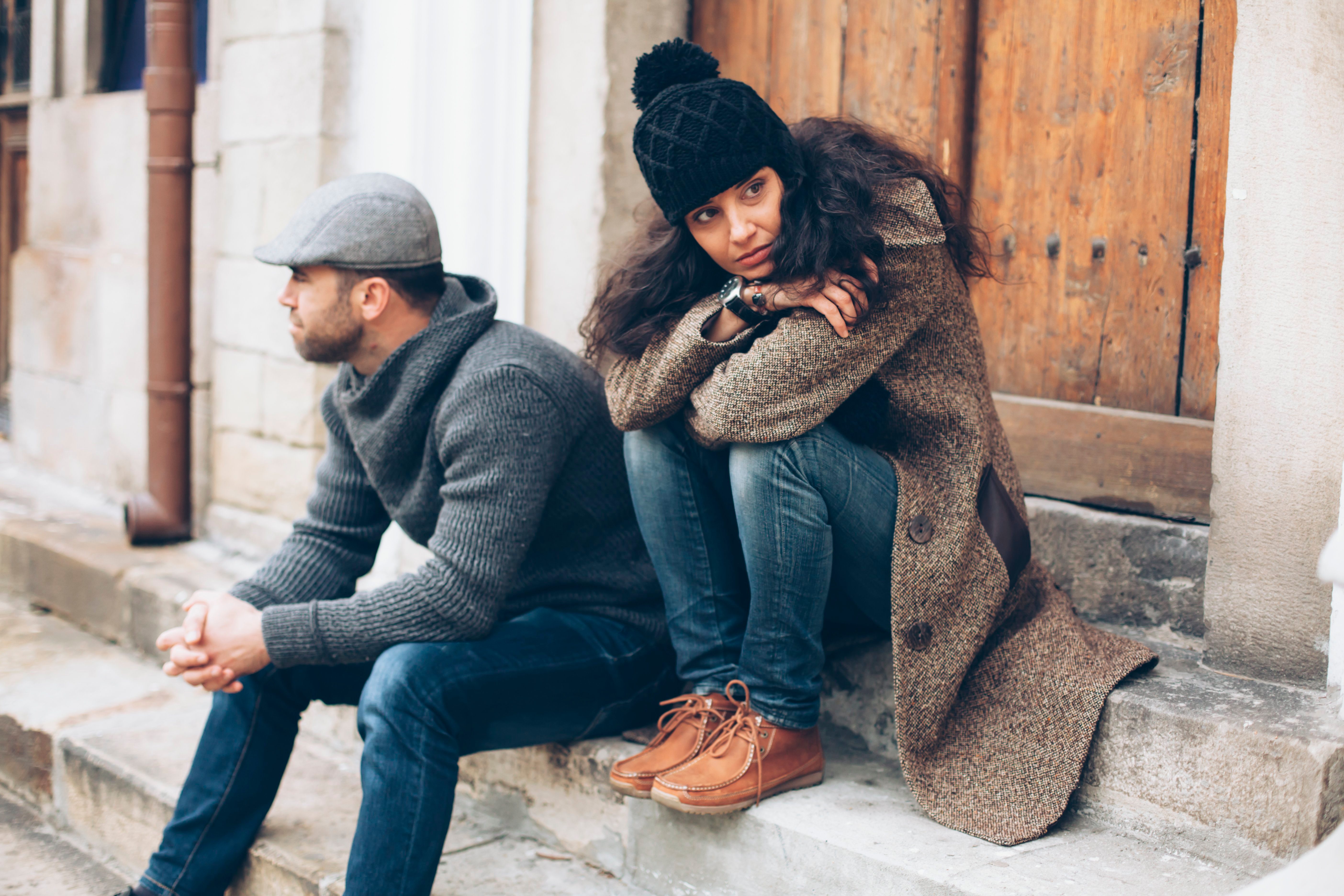 Young couple having relationship difficulties. Man and woman sitting on stairs in front of a closed wooden door. Wears warm clothes - jeans, sweater, coat, hat and scarf. Both looking in different directions. On background building facade, wooden front door, rainwater gutter and windows.