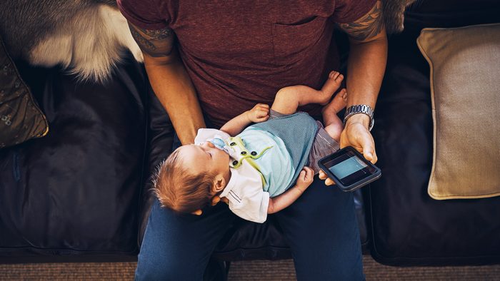 young man using a mobile phone while holding his newborn baby boy at home