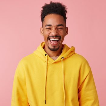 Most funny jokes - Young happy smiling african american guy in yellow hoodie, heard a very funny joke and laughed, standing on a pink background with eyes closed