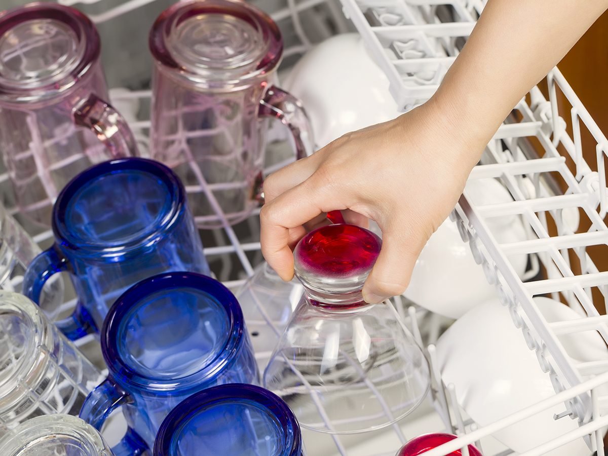 Horizontal photo of female hand putting in glassware to be washed by dishwasher with blue, red, clear, pink glasses and white bowls in background