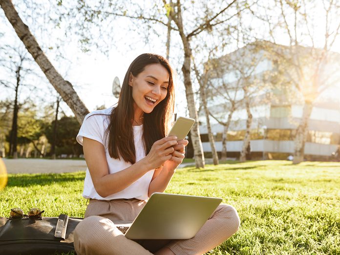 Photo of happy woman sitting on grass outdoors using laptop computer and mobile phone.