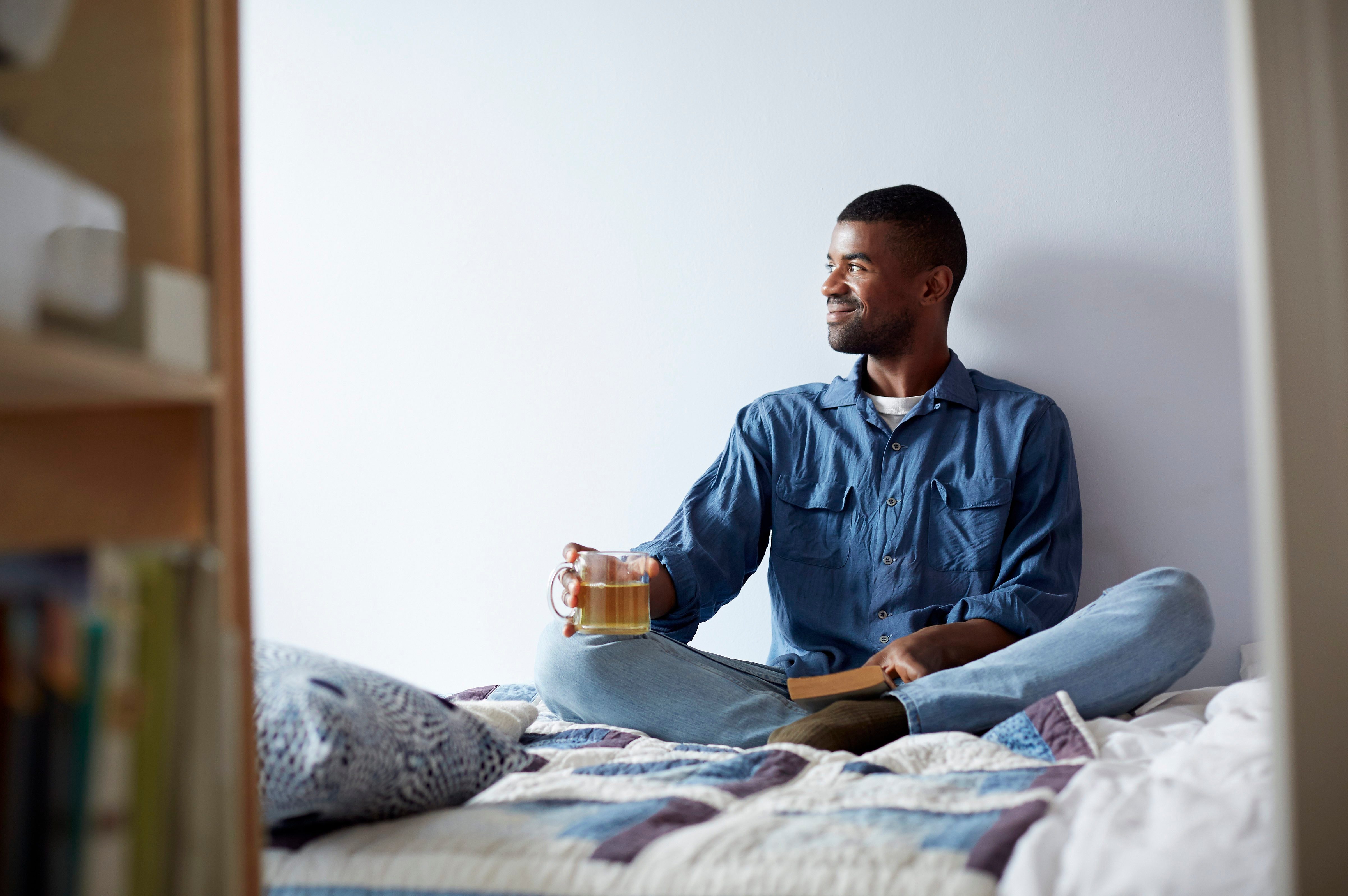Black man drinking tea and reading on bed
