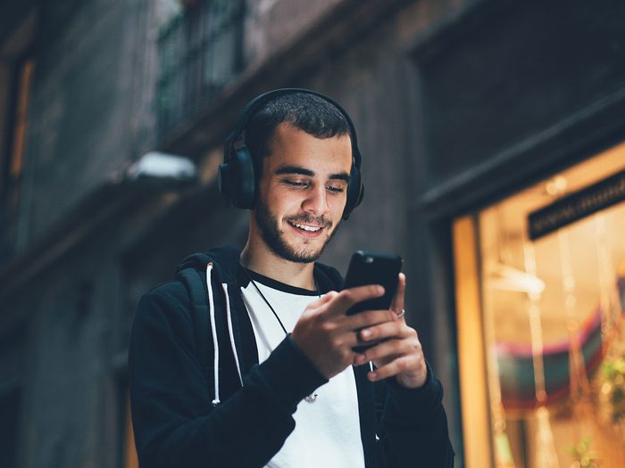Signs of hearing loss - Handsome young man in black hoodie wears big wireless music headphones and changes audio tracks on smartphone, he smiles when looks at phone screen