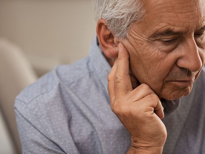 Side view of senior man with symptom of hearing loss. Mature man sitting on couch with fingers near ear suffering pain.