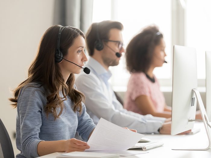 Work jokes - Diverse call center workers sitting in row, focus on customer service member girl in headset looks at pc screen hold paper talk with potential client provide insurance information sell company product