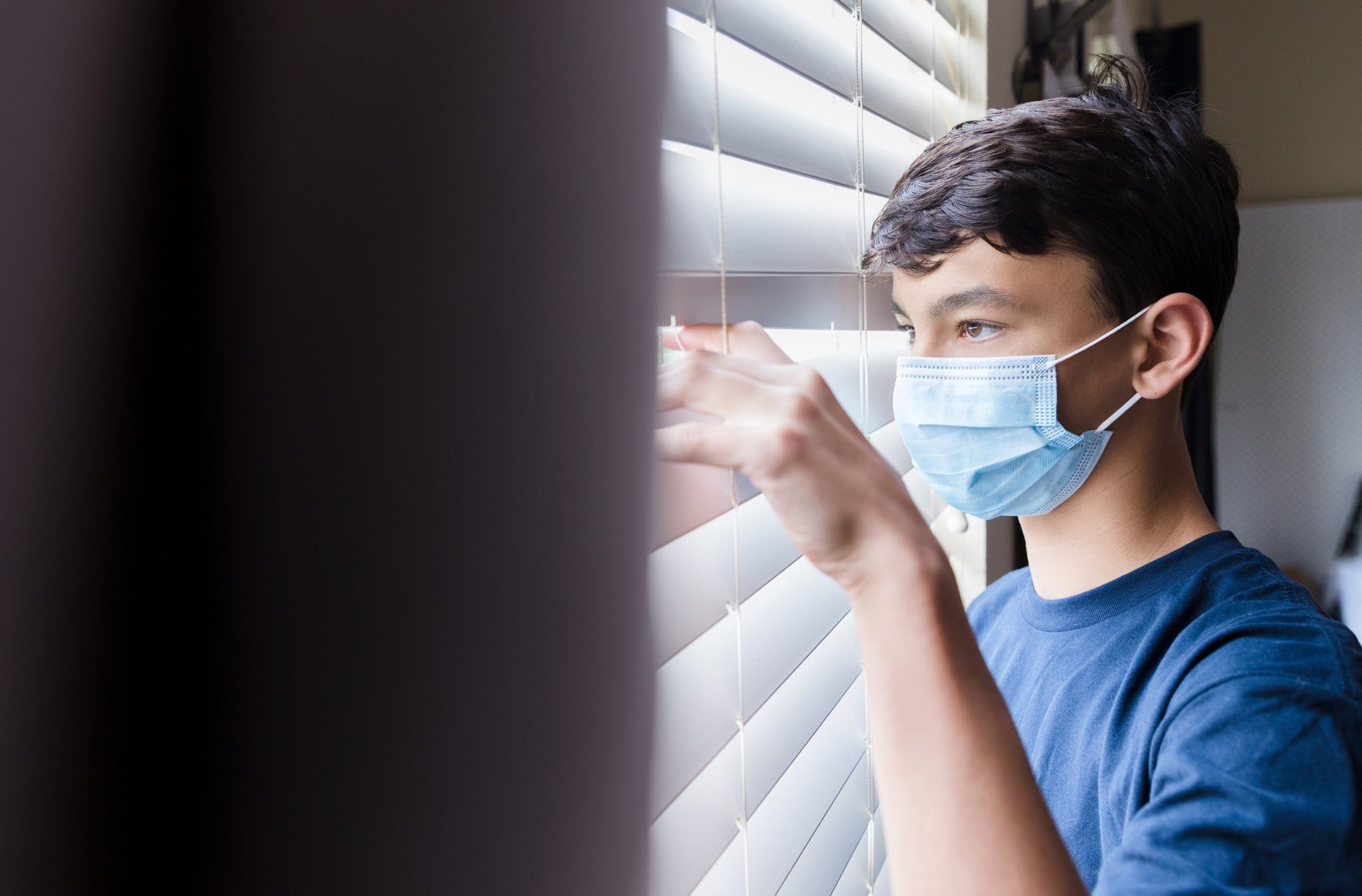 During COVID-19, quarantined boy wearing mask looks through blinds