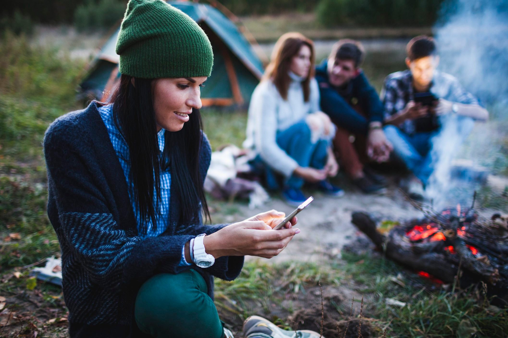 Young woman using mobile phone while friends sitting by bonfire at campsite