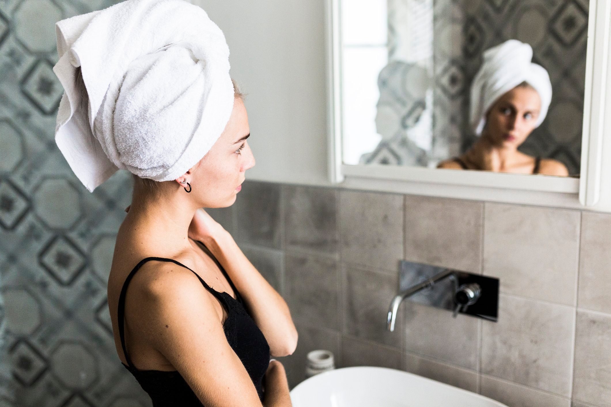 young woman with hair wrapped in towel looking at herself in the bathroom mirror