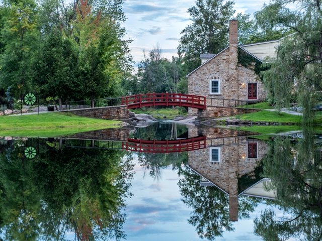 Day trips from Ottawa - In Stewart Park, a view of the red footbridge and old stone house surrounded by trees, as they are reflected in the perfectly still pond