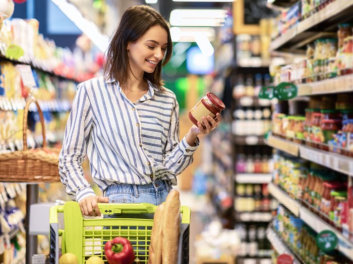 Portrait Of Smiling Woman With Shopping Cart In Supermarket Buying Groceries Food Walking Along The Aisle And Shelves In Grocery Store, Holding Glass Jar Of Sauce, Choosing Healthy Products In Mall