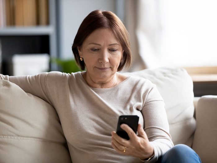Worried middle aged woman sitting on couch, looking at mobile phone screen.