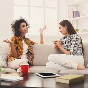 Rude conversation habits - Two happy young female friends with coffee cups conversing in living room at home, chatting about their life and relations, gossip and slumber party concept, copy space