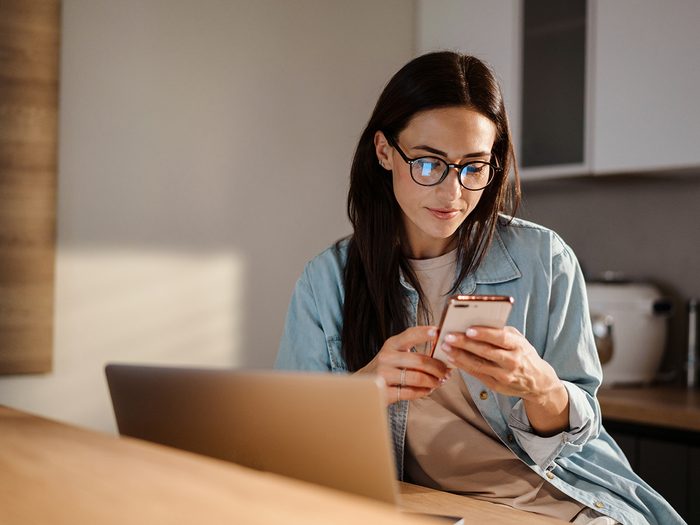 Serious charming woman using smartphone while working with laptop at home