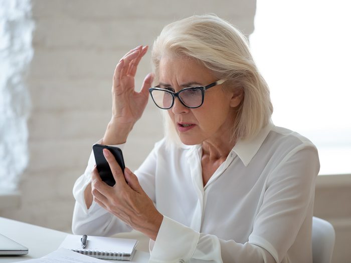 Confused senior businesswoman sit at office desk hold cellphone experience internet connection problem, frustrated aged woman worker feel disappointed having smartphone breakdown or virus attack