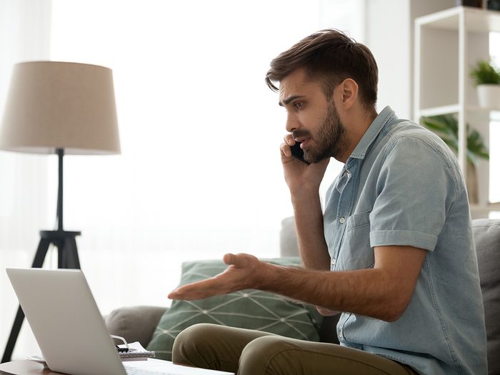 Confused male having computer problems talk on mobile with customer support, frustrated millennial man speak on smartphone managing computer malfunction working online from home