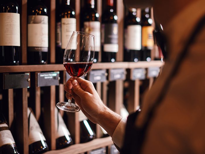 worst foods for brain - Bartender or male cavist standing near the shelves of wine bottles holds a glass of wine, looks at tint and smells flavor of wine in glass.