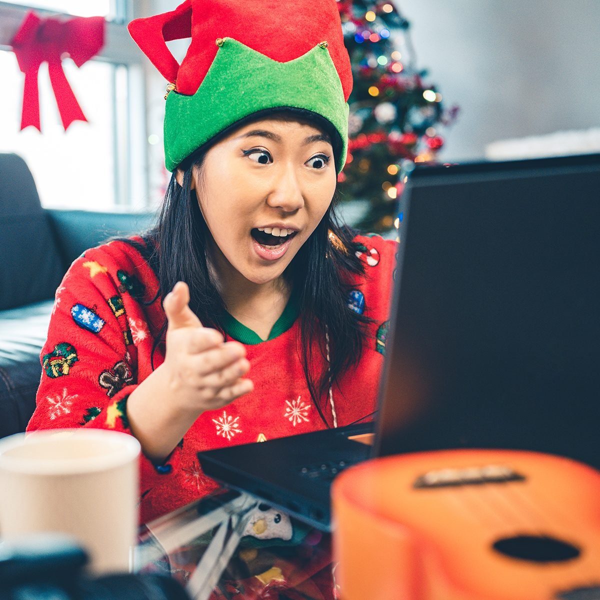 Millennial Christmas party in season costumes. Interior of modern apartment with Christmas decorations. Portrait of a young Chinese woman looking at laptop