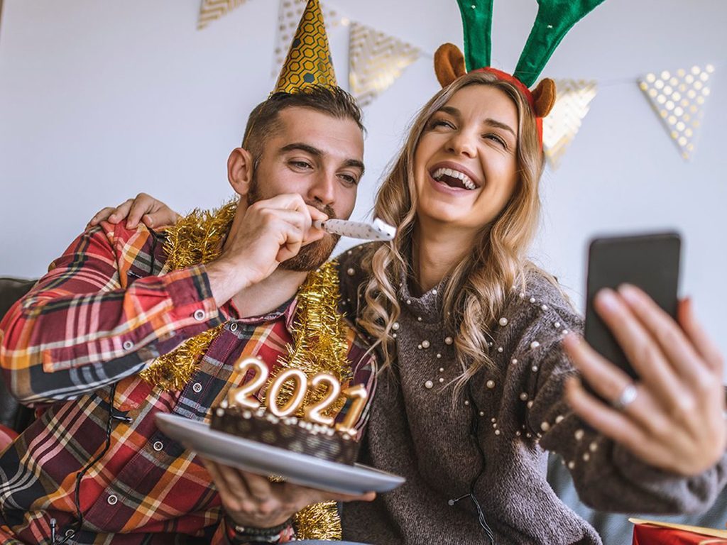 Couple celebrating Christmas at home, holding chocolate cake with lit candles shaped as numbers 2021 and having a video call with friends