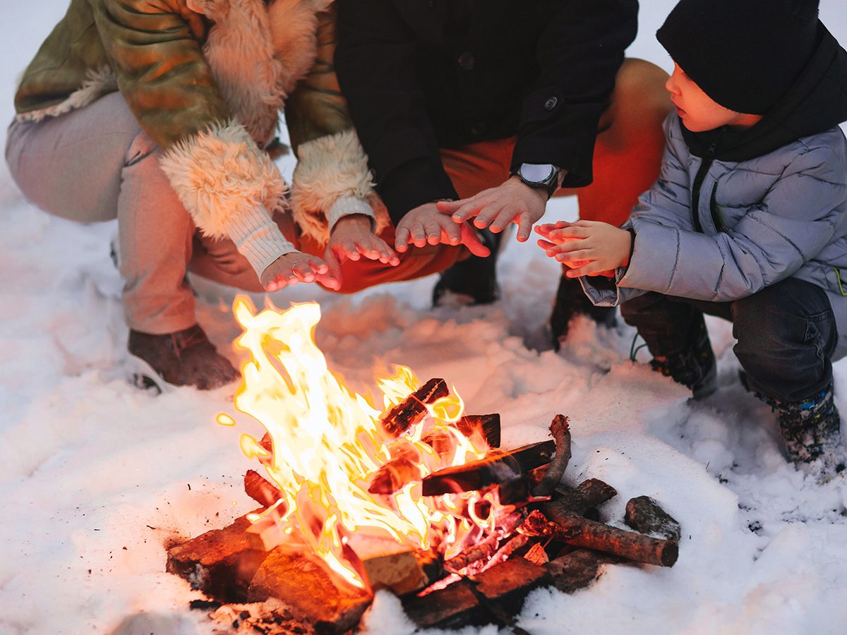 how your body deals with freezing weather - Happy family of three, mother, father and son, in warm clothes, sitting close to each other and trying to warm up by fire outside in cold snowy winter weather, against snowbound forest background