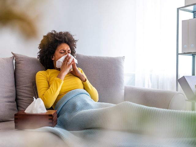 Picture showing sick woman sneezing at home. Young sick woman sneezing in tissue sweating from flu fever. Sick woman catch cold. Sneezing with handkerchief, coughing, got flu, having runny nose.