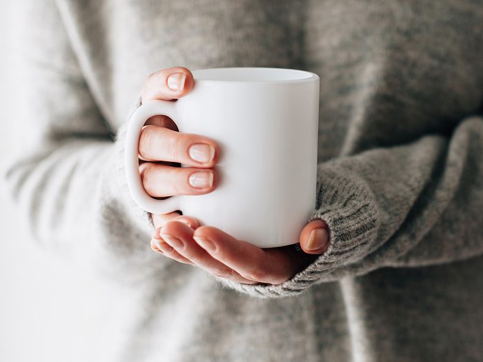 Do You Need to Wash Your Coffee Mug Every Day - Closeup of female hands with a mug of beverage. Beautiful girl in grey sweater holding cup of tea or coffee in the morning sunlight