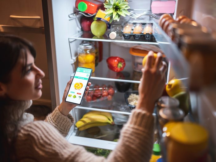 Keep your food from spoiling - Beautiful young woman standing next to an opened refrigerator door, holding a smart phone and ordering fresh fruit and vegetables online for home delivery